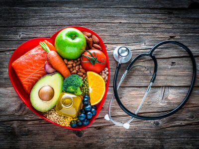 Healthy eating: fresh multicolored foods to help lower cholesterol levels and for heart care shot on wooden table. The composition includes oily fish like salmon. Beans like Pinto beans, soybeans and brown lentils. Vegetables like garlic, carrot, avocado, broccoli and tomato. Fruits like apple, orange and berries. Nuts like almonds. Olive oil. The food is arranged in a red heart shape tray and a stethoscope is beside it. High resolution 42Mp studio digital capture taken with SONY A7rII and Zeiss Batis 40mm F2.0 CF lens
